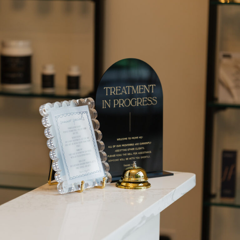 Prime MD medical spa reception area in Rolesville, NC featuring elegant ‘Treatment in Progress’ sign, gold service bell, and framed summer specials on a white marble counter-modern medspa offering Botox, filler, laser treatments, facials, and wellness services.
